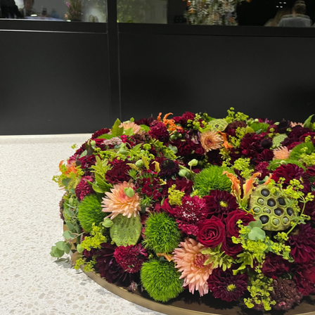 Eine kreisförmige Blumenarrangement mit einer Mischung aus roten, grünen und orangefarbenen Blumen. Es steht auf einer weißen Oberfläche vor einer schwarzen Wand mit einer Glasscheibe.