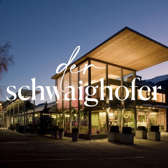 Beleuchtetes modernes Gebäude mit Glaswänden, einem Baum und einem Namen in weißer Schrift. Mehrere Topfpflanzen stehen davor.