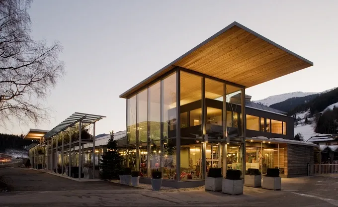 Ein modernes Gebäude mit Glaswänden und einem Holzdach. Es hat eine große Glaswand und einen Balkon. Es ist von Pflanzen und Bäumen umgeben.