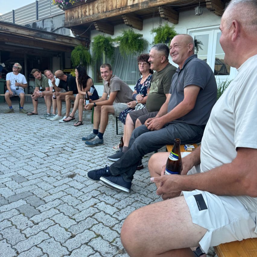 Eine Gruppe von Menschen sitzt auf einem gepflasterten Weg, einige halten Flaschen, lächeln und schauen zur Kamera. Hinter ihnen ein Gebäude mit Pflanzen und einem Balkon.