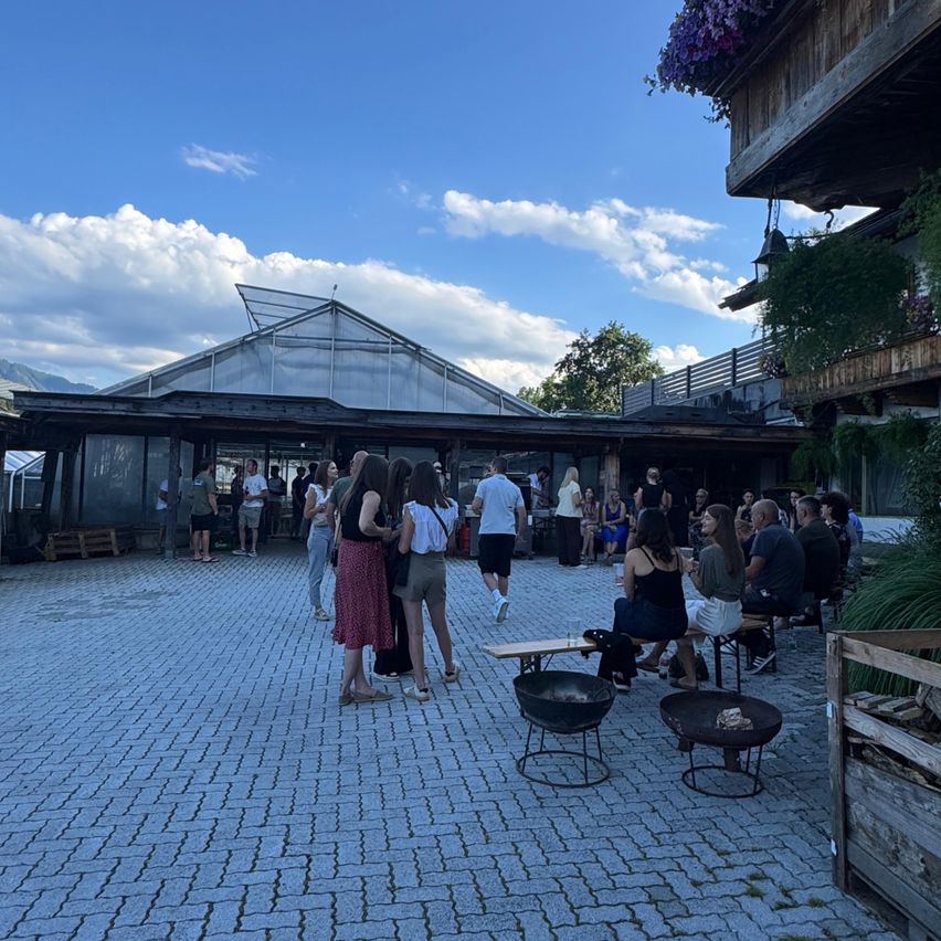 Eine Gruppe von Menschen versammelt sich vor einem Gebäude mit Glasdach, einige sitzen auf Bänken und andere stehen, unter einem blauen Himmel mit Wolken.
