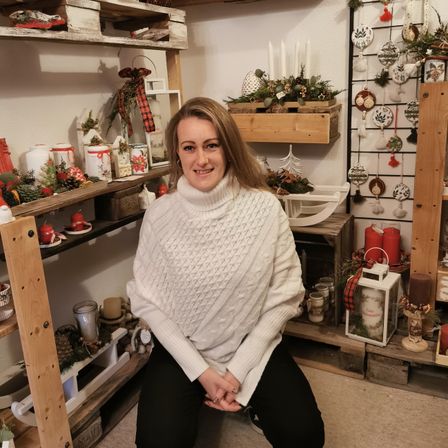 Eine Frau sitzt in einem mit Weihnachtsdekorationen gefüllten Raum. Sie trägt ein weißes Rollkragenshirt und schwarze Hosen. Regale halten Kerzen, Ornamente und Pflanzen.