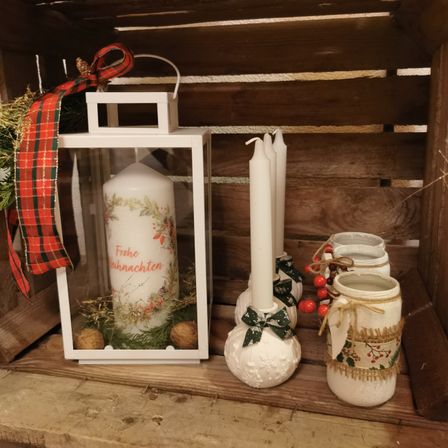 Eine Weihnachtskerze mit einem festlichen Band befindet sich in einer Laterne. Drei weiße Kerzen und Gläser sind neben ihr auf einem Holzregal angeordnet.