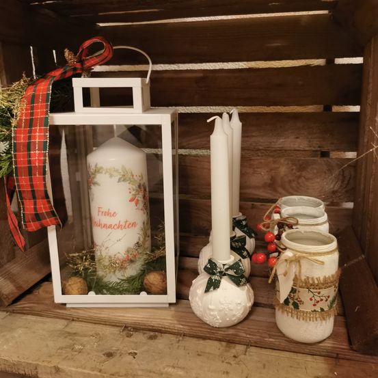 Eine Weihnachtskerze mit einem festlichen Band befindet sich in einer Laterne. Drei weiße Kerzen und Gläser sind neben ihr auf einem Holzregal angeordnet.