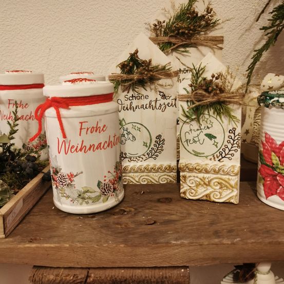 Ein Holzregal hält Keramikgläser und Schachteln mit Weihnachtsdekoration, einschließlich roter Bänder und grüner Pflanzen. Ein Glas ist mit 'Frohe Weihnachten' in roten Buchstaben beschriftet. Hinter den Gläsern befinden sich festliche Schachteln, die mit Juteschnur gebunden sind.