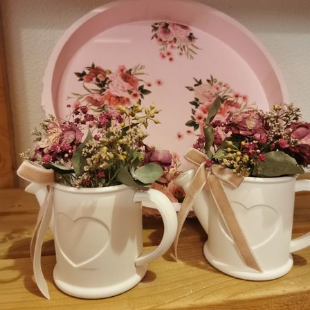 Zwei weiße Keramik-Gießkannen mit Herz-Designs auf einem Holzregal, geschmückt mit getrockneten Blumen und Bändern. Dahinter befindet sich ein rosa Teller mit Blumenmustern.
