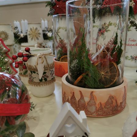 Auf einem Tisch sind verschiedene Weihnachtsdekorationen zu sehen. Zentral steht eine hohe Glasvase mit Tannenzapfen, Grünpflanzen und Orangenscheiben. Andere Gegenstände umfassen ein Glas mit einem festlichen Band, eine kleine gnomenförmige Basis und eine Glaskugel mit roten Blumen. Im Hintergrund befinden sich Kerzen und andere Ornamente.