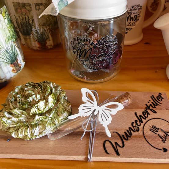 Ein Glas mit Deckel enthält eine Blume und einen Schmetterling auf einem Holzbrett. Das Glas ist mit dem Text 'MAMA Super' beschriftet. Das Brett hat die Worte 'Wuenscheruefllen'.