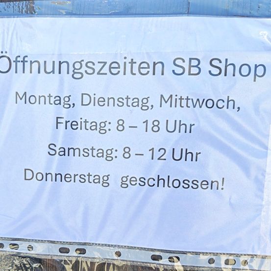 Das Schild zeigt die Öffnungszeiten für den SB Shop. Geöffnet Montag, Dienstag, Mittwoch, Freitag von 8-18. Samstags ist es 8-12. Sonntags geschlossen.
