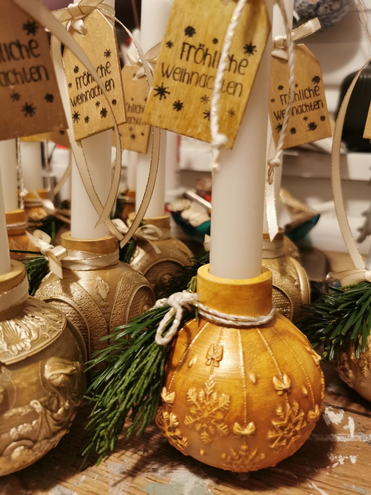 Eine Ausstellung goldener Kerzen mit aufwendigen Designs und grünen Zweigen, geschmückt mit Etiketten, die 'Frohe Weihnachten' lesen. Jede Kerze ist in ein weißes Band gewickelt.