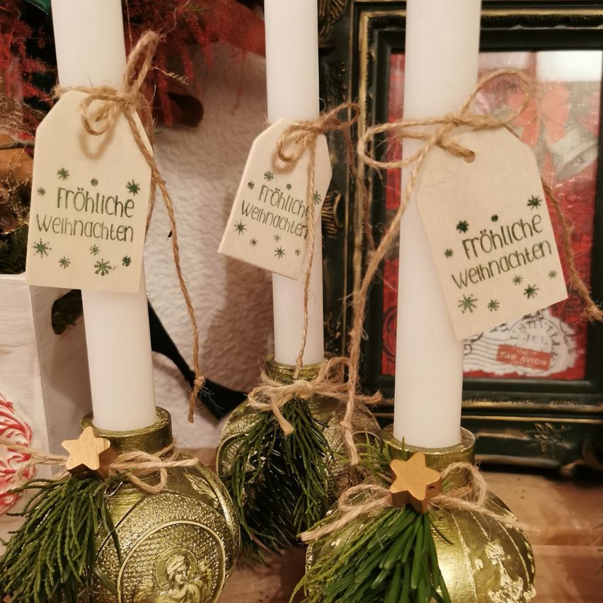 Drei goldene und grüne dekorierte Weihnachtskerzen mit weißen Kerzen und einem Schild, auf dem 'Frohe Weihnachten' steht, stehen auf einer Holzoberfläche. Dahinter befindet sich ein Bilderrahmen mit rotem Hintergrund.