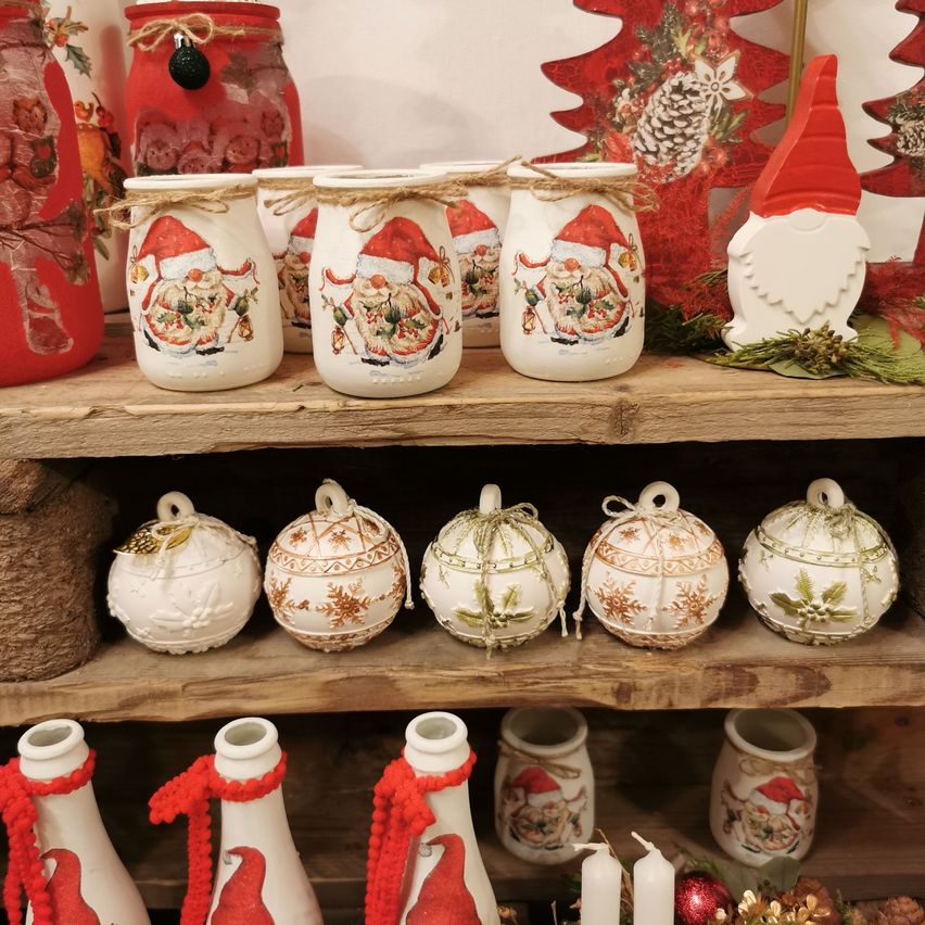 Ein Holzregal zeigt festliche Gegenstände, darunter Gläser mit Weihnachtsmann-Designs, Gnom-Figuren und Weihnachtskugeln. Jedes Glas ist mit rustikalem Tau umwickelt, und die Ornamente sind kunstvoll verziert. Auf dem unteren Regal befinden sich auch rote und weiße Flaschen mit Weihnachtsmann-Illustrationen und Kerzen.