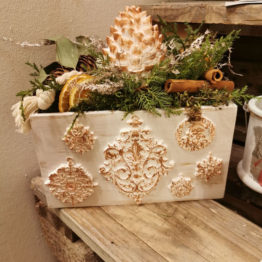 Ein weißer Pflanzkasten mit kunstvollen goldenen Mustern, gefüllt mit einer Tannenzapfen, grünem Laub, Orangenscheiben und Zimtstangen. Er steht auf einer Holzoberfläche.