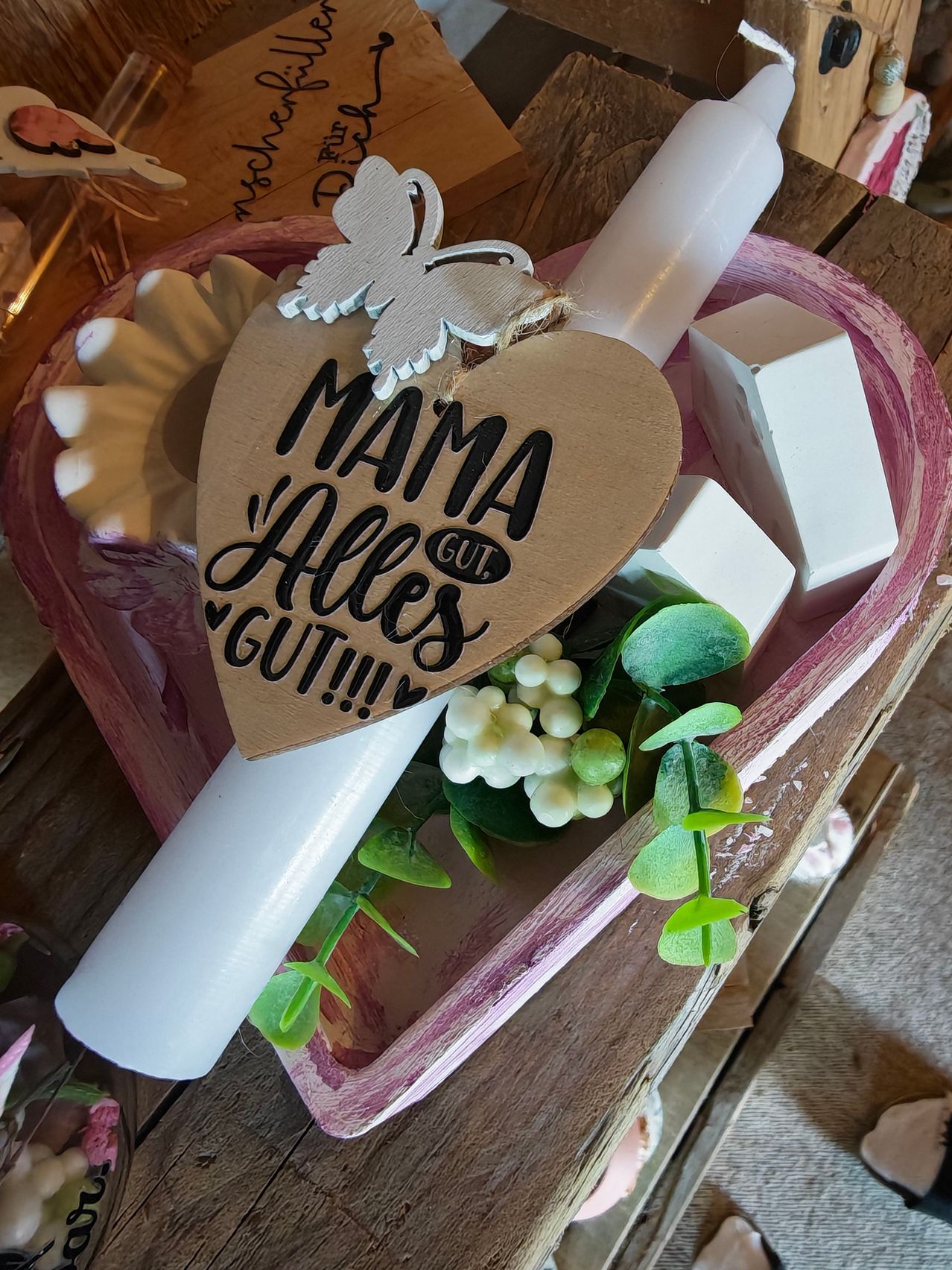 Ein herzförmiges Holzschild mit der Aufschrift 'MAMA GUT ALLE GUT!!!' hängt an einer weißen Kerze in einem rosa Korb mit Blumen und Seifen.