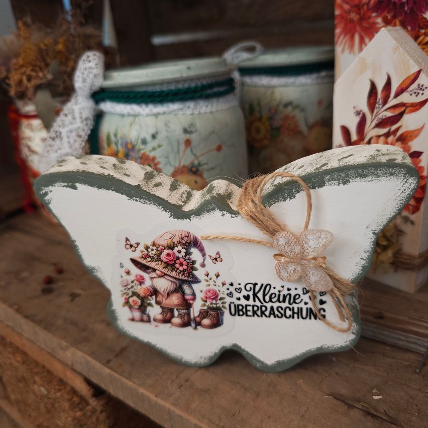 Eine dekorative hölzerne Schmetterlingsfigur mit einem Gnom-Design, mit einem Seil gebunden, und den Worten 'Kleine Uberraschung' darauf. Dahinter befinden sich Gläser mit Blumendesigns.
