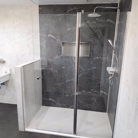 Eine moderne Duschkabine mit einer Glastür und einer Duschbrause an einem metallischen Arm, neben einem weißen Waschbecken. Die Wände und der Boden sind mit grauem Marmor gefliest.