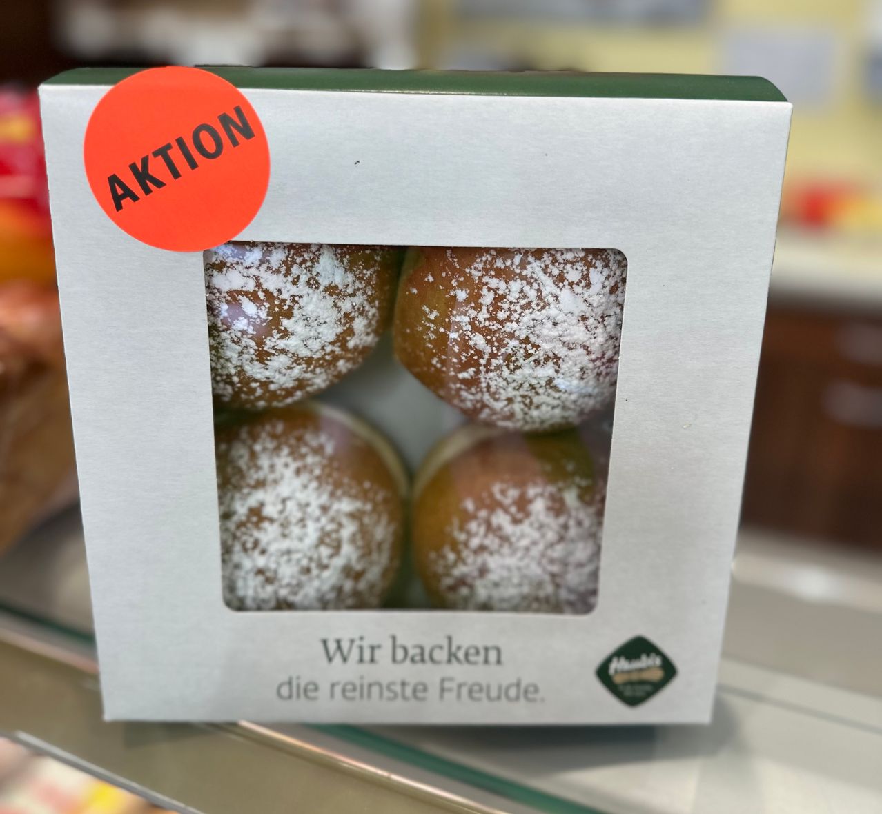 Eine weiße Schachtel mit grünem Deckel enthält vier runde Donuts, die mit Puderzucker bedeckt sind. Ein roter Aufkleber mit dem Wort 'AKTION' befindet sich oben links.