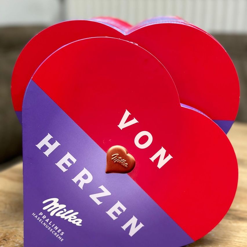 A heart-shaped box of chocolates labeled 'Von Herzen' with a brown chocolate in the middle, resting on a wooden surface. The box has red and purple colors.