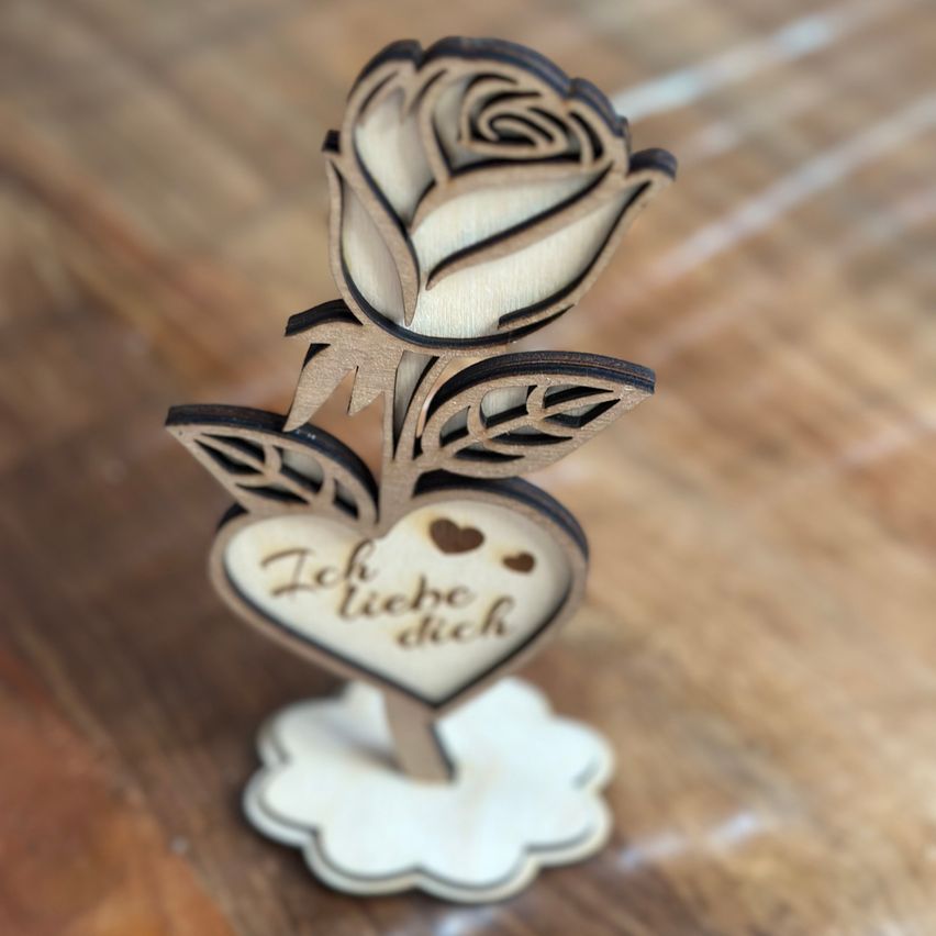 A wooden heart with two roses on top, and a cloud base, has a message written on it that reads 'I love you'. The object is placed on a wooden surface.