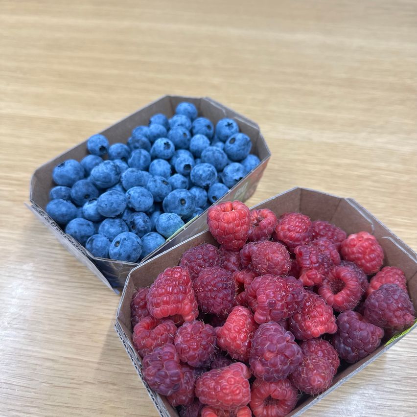 Zwei Pappkartons auf einer Holzoberfläche enthalten Blaubeeren und Himbeeren. Die Blaubeeren sind tiefblau und die Himbeeren sind leuchtend rot.