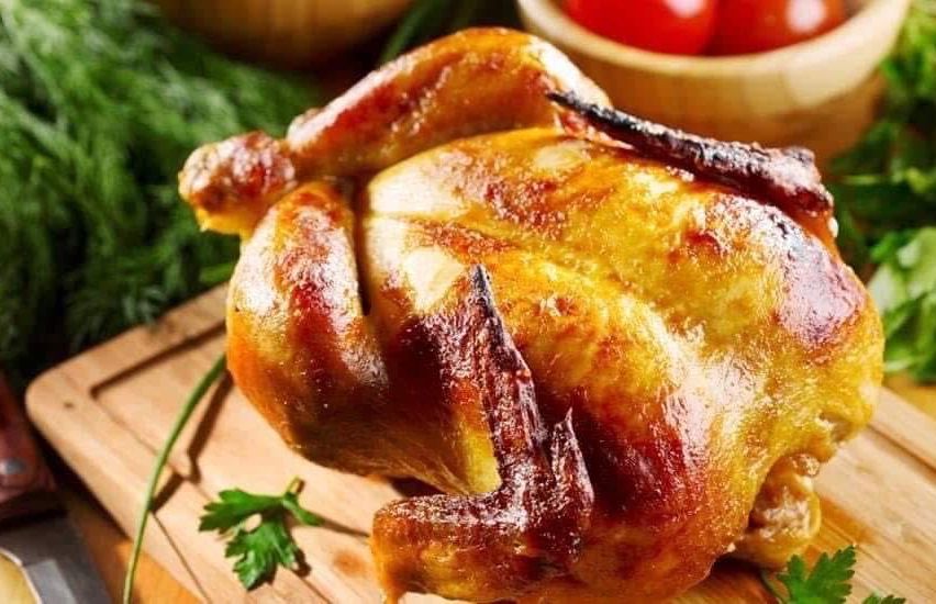 A roasted chicken sits on a wooden cutting board with herbs, ready to be served.