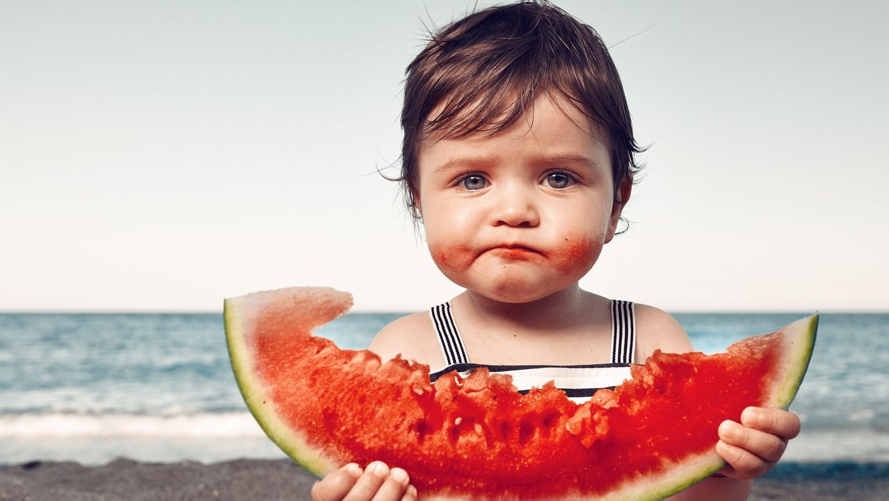 Ein junges Kind hält eine große Scheibe Wassermelone mit einem verschwitzten Gesicht und steht auf einem Strand mit einer ruhigen Ozean im Hintergrund.