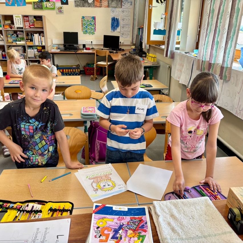Drei Kinder arbeiten in einem Klassenzimmer an einer Malseite. Sie haben Buntstifte und Malbücher.