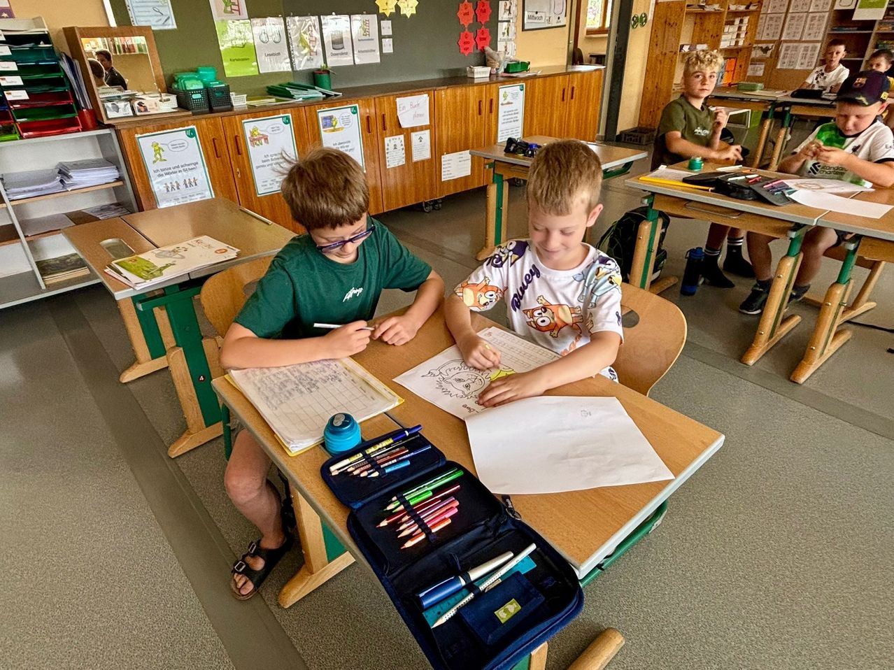 Zwei Jungen sitzen an einem Schreibtisch in einem Klassenzimmer, schreiben und zeichnen. Sie sind von Schulmaterialien umgeben. Ein anderer Schüler sitzt hinter ihnen an einem Tisch.