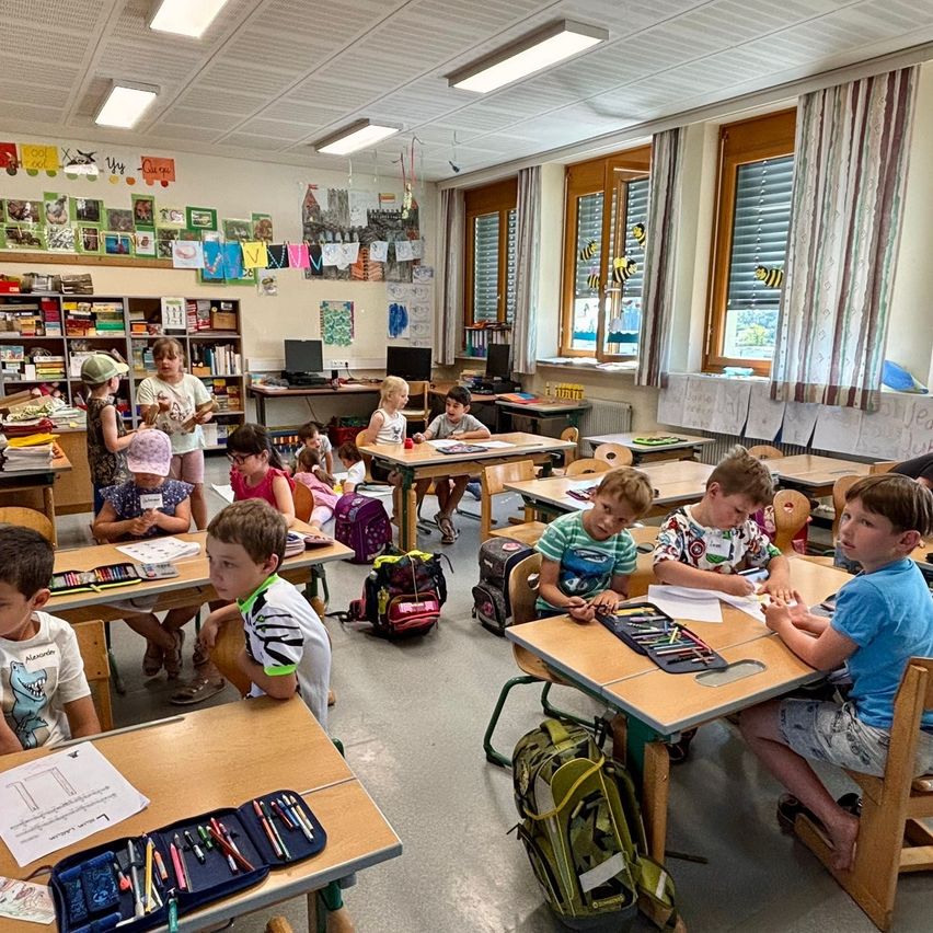 Ein Klassenzimmer voller Kinder, die an Tischen sitzen. Einige benutzen Stifte, und es gibt Bücher und Taschen auf den Tischen. Der Raum hat Regale mit Büchern, Fenster mit Jalousien und verschiedene Poster an den Wänden.