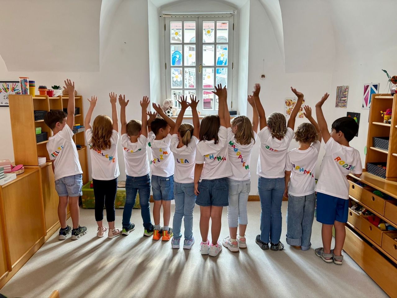 Eine Gruppe von Kindern steht in einem Kreis mit erhobenen Händen in einem hellen Raum mit weißen Wänden und Boden. Sie tragen weiße T-Shirts mit buntem Text. Ein Fenster mit blumengeschmücktem Glas ist hinter ihnen.