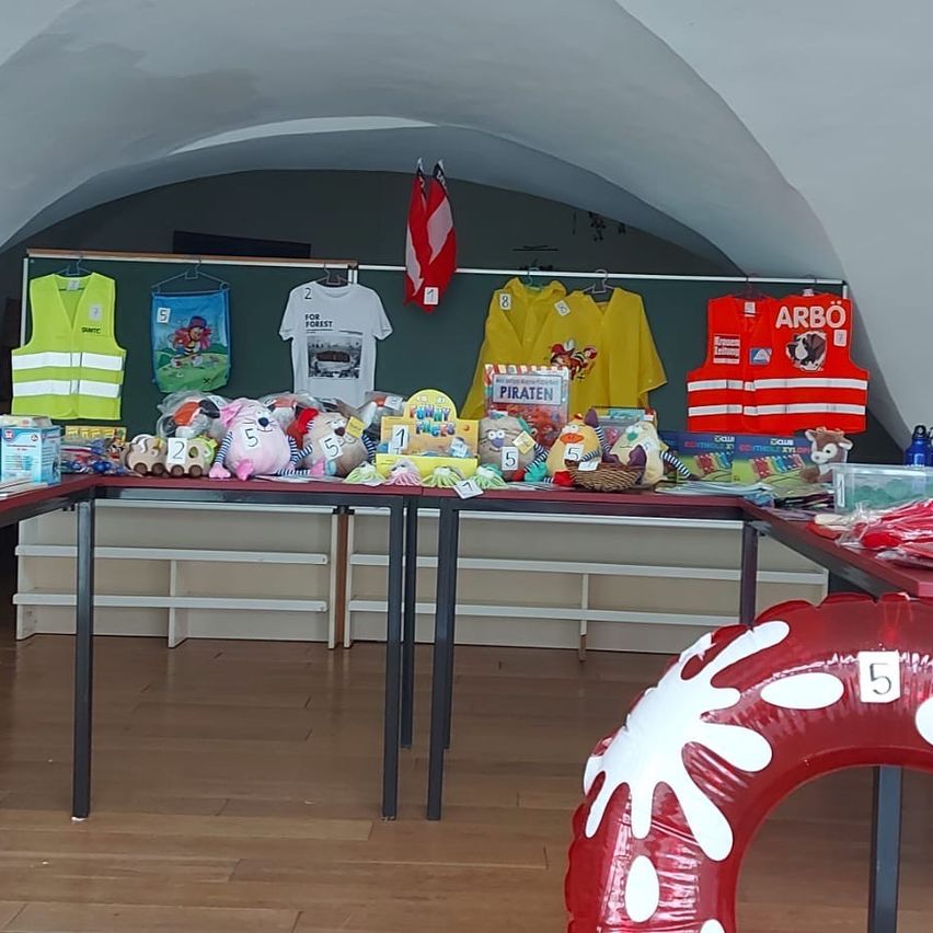 Ein Raum mit verschiedenen Gegenständen auf Tischen, darunter ein roter Rettungsweste mit der Aufschrift Arbo, ein gelber Regenmantel, eine gelbe Weste und ein weißes T-Shirt. Es gibt auch Spielzeug, Bücher und einen roten Ballon auf dem Boden.
