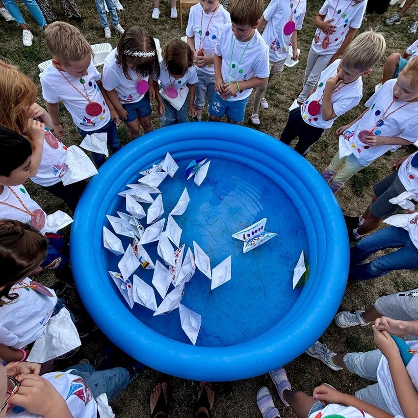 Eine Gruppe von Kindern steht um einen blauen Pool mit Papierbooten herum, alle tragen weiße T-Shirts mit bunten Designs. Einige halten Papierboote und schauen in den Pool.