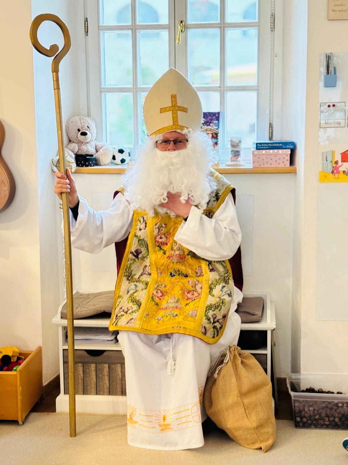 Ein Mann, verkleidet als Bischof, hält einen Stab in einem Raum mit Fenster, Spielzeug, Büchern und einem Beutel.