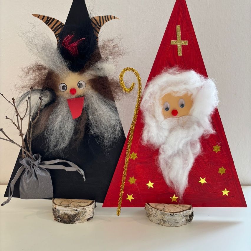 Zwei handgefertigte Weihnachtsdekorationen auf einer weißen Oberfläche. Die linke ist eine Hexe mit schwarzem Hut und roter Zunge. Die rechte ist eine Weihnachtsmannfigur mit rotem Hut, weißem Bart und einem goldenen Kreuz obenauf.