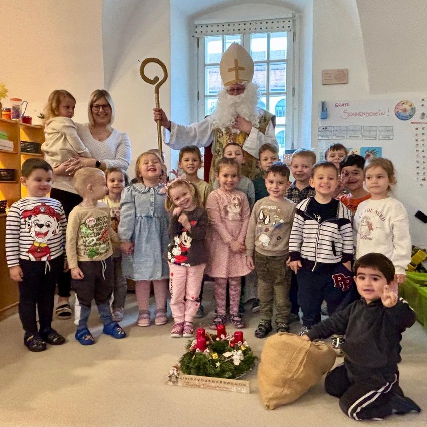 Eine Gruppe von Kindern und Erwachsenen in einem Raum, einige stehend und andere sitzend, mit einer als Weihnachtsmann verkleideten Person in der Mitte. Einige Kerzen stehen auf einem Tisch vor ihnen.