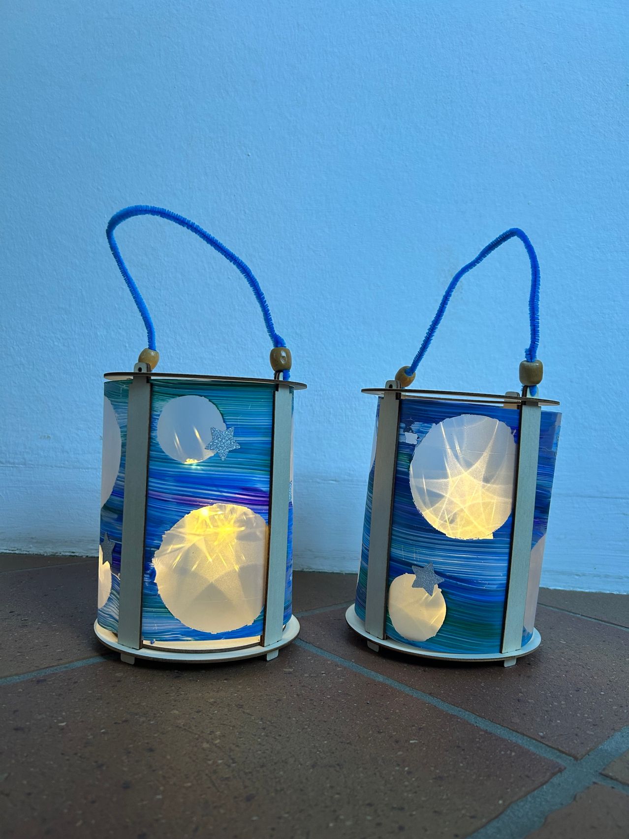 Zwei zylindrische Laternen mit blauem Hintergrund und weißen kreisförmigen Designs, von innen beleuchtet. Sie haben blaue Griffe und Sockel.