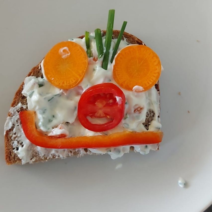 Ein Stück Brot mit einer cremigen Aufstrich und Gemüsegesicht, bestehend aus Karotten, einer Tomate und einer roten Paprika.