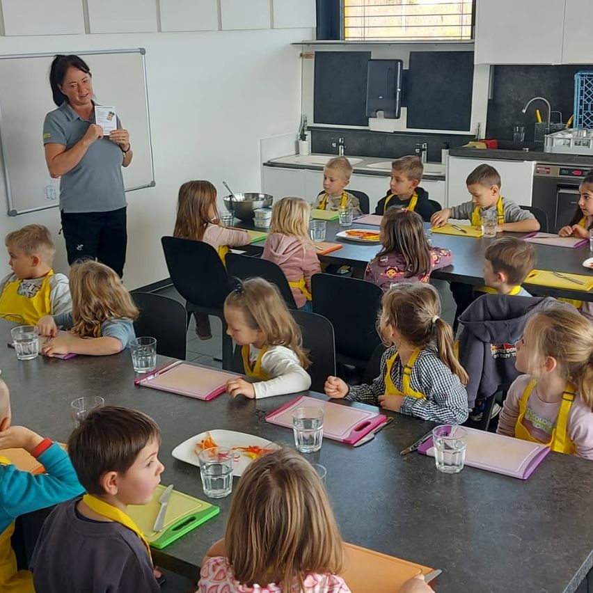Eine Gruppe von Kindern sitzt an einem langen Tisch in einer Küche und bereitet Essen zu. Eine Frau steht an einer Tafel und hält ein Stück Papier in der Hand. Verschiedene Küchenutensilien, einschließlich Schüsseln, Teller und Besteck, befinden sich auf dem Tisch.