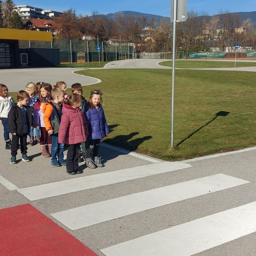 Eine Gruppe von Kindern steht auf einem Bürgersteig, mit einem Grasfeld und einem Pfosten hinter ihnen. Die Sonne wirft Schatten auf den Boden. Gebäude und Berge sind in der Ferne sichtbar.