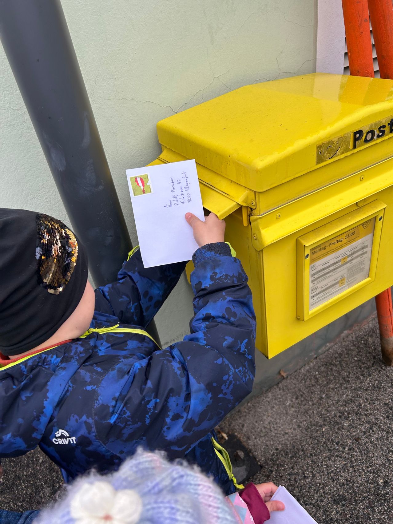A young child wearing a blue winter jacket and a black beanie puts a white envelope into a yellow postal box.