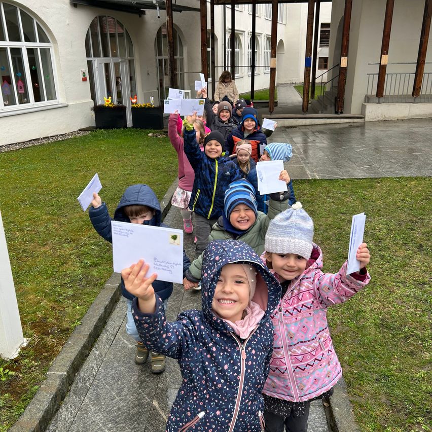 Children in rain jackets hold letters and smile, lined up outside a building with arched windows.