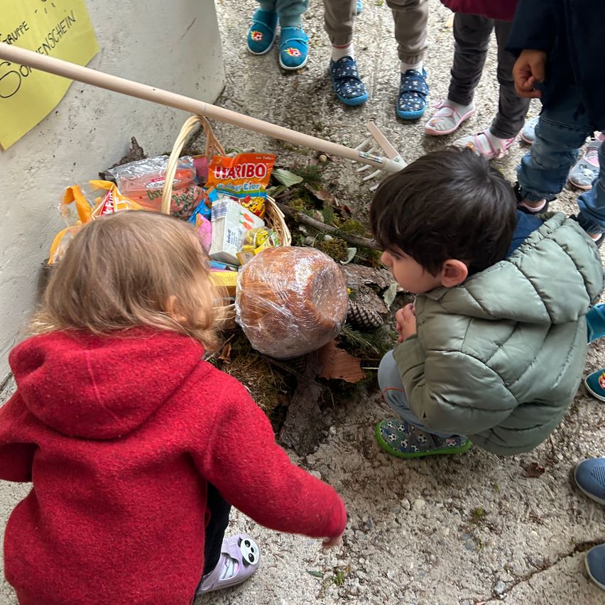 A group of children inspects a basket of snacks and a loaf of bread on the ground. Nearby, others stand with slippers and a rake.