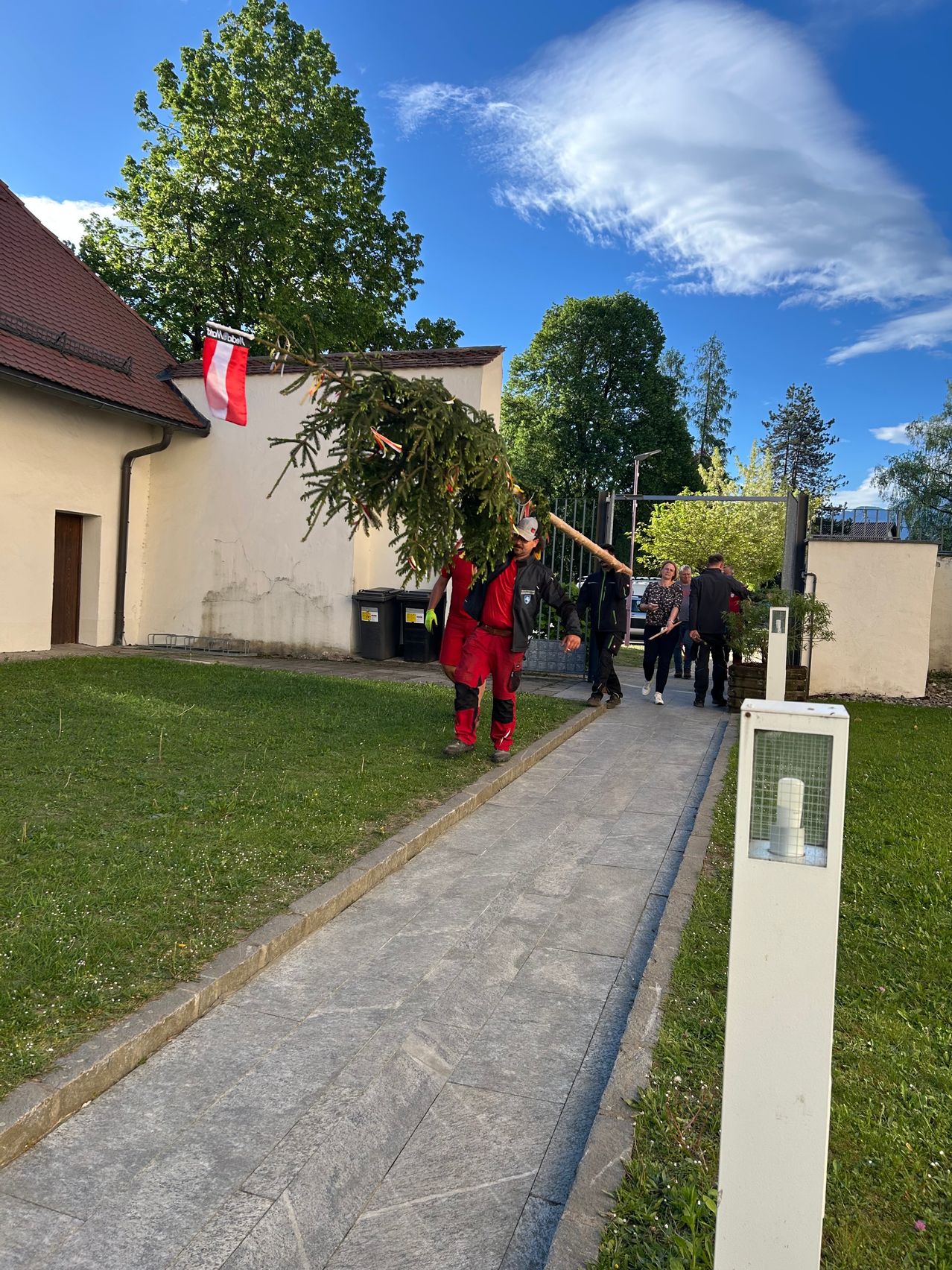A person carrying a fir tree on a pole and walking on the sidewalk outside the building with a flag above.