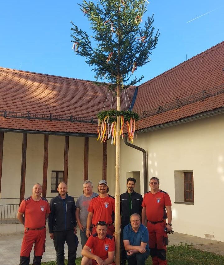 Eight men in red and blue uniforms stand around a decorated Christmas tree outside a building with a red roof.
