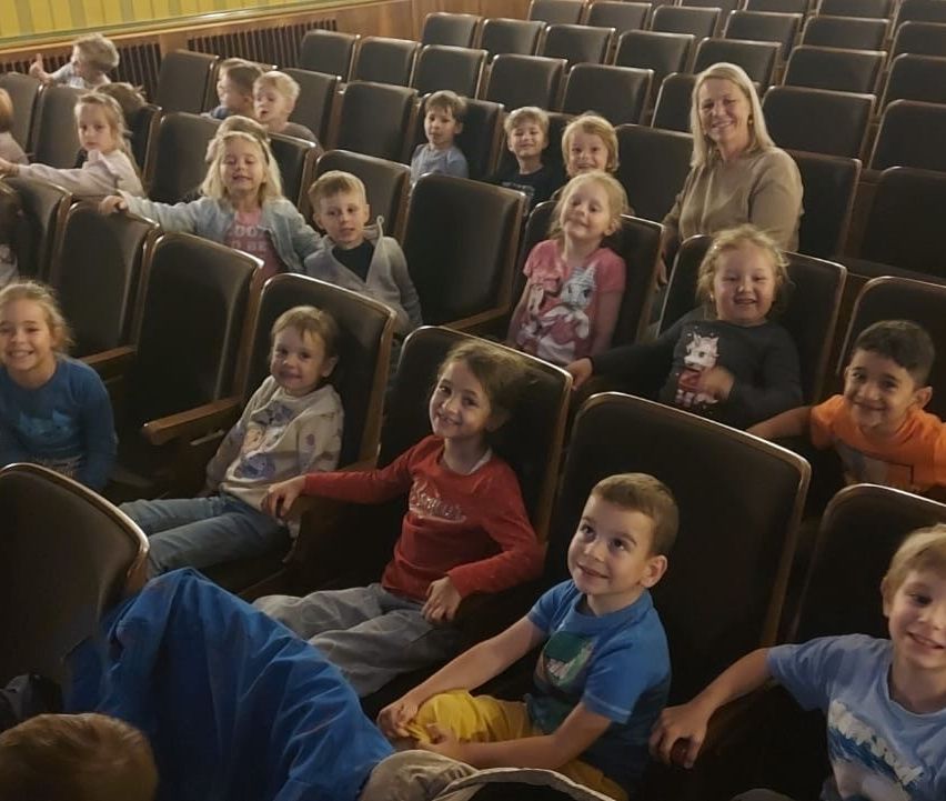 A group of children and adults are seated in a theater, all smiling and looking forward, with a woman in front smiling at the camera.