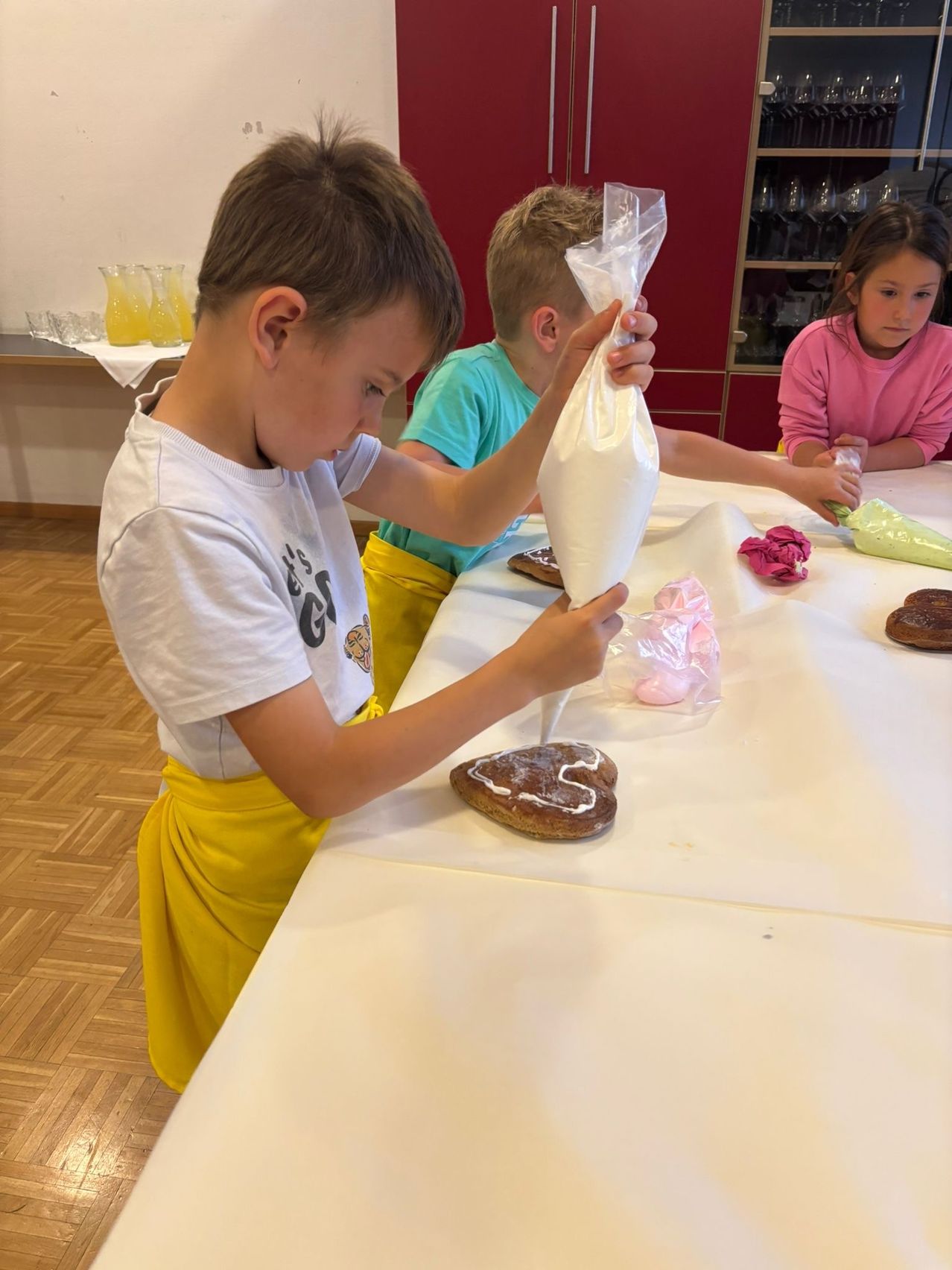 Three kids are decorating cookies with frosting. One boy is using a piping bag, another is holding a bag of frosting, and a girl is decorating cookies with icing.