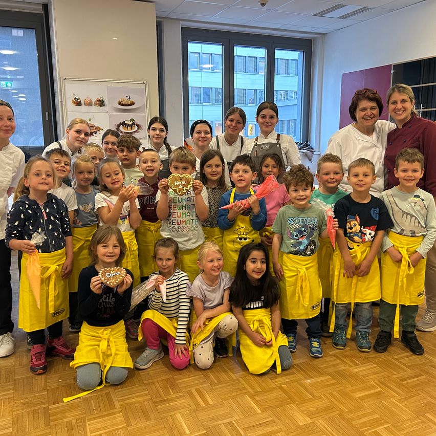 A group of children and adults in a room with yellow aprons, holding heart-shaped cookies, posing for a photo. There is a whiteboard with food images and two women standing in front of it.