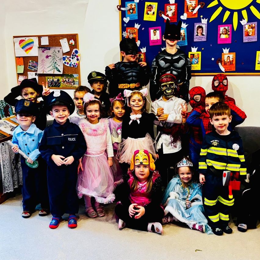 A group of children dressed in various costumes, including Batman, Spiderman, police, and firefighters, pose for a photo in a room with a bulletin board in the background.