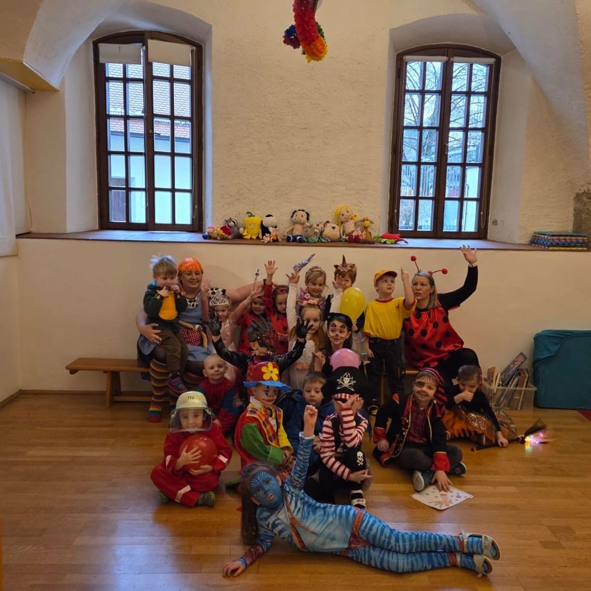 A group of children dressed in various costumes are sitting and lying on a wooden floor in a room with white walls and windows.