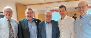 Fünf Männer stehen in einem Raum und posieren für ein Foto. Sie alle lächeln und tragen formelle Kleidung. Hinter ihnen steht ein weißer Tisch und eine Person steht in der Nähe der Wand.